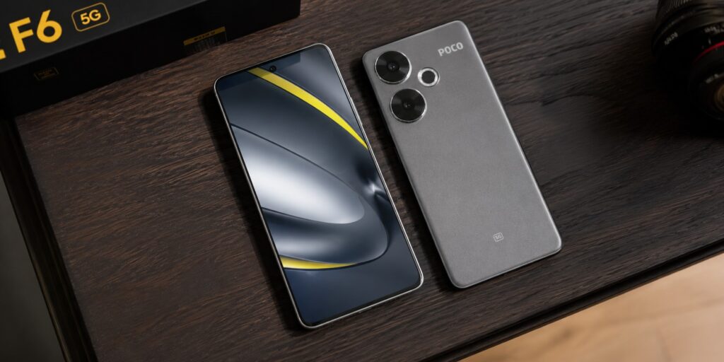 Poco F6 5G smartphone in Titanium matte finish, displaying its large dual camera rings and flat AMOLED screen, positioned on a wooden desk.