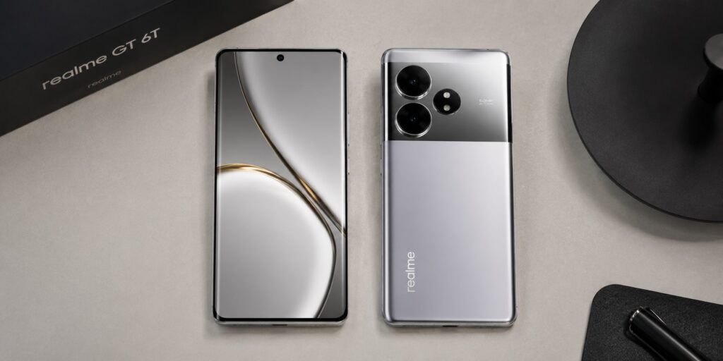 Realme GT 6T smartphone in Fluid Silver showing its dual-tone mirror back panel and 1.5K curved LTPO AMOLED display on a desk.